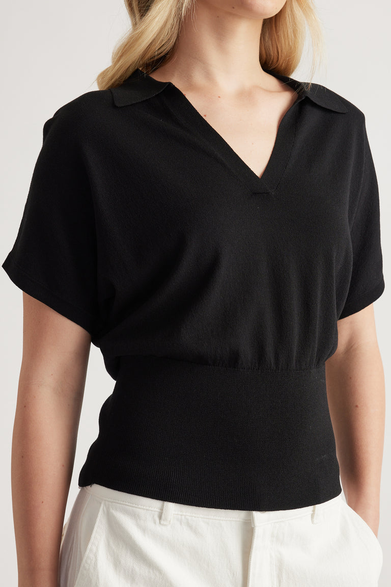 Woman wearing a black short sleeve collared knit top with a cinched waist and white pants