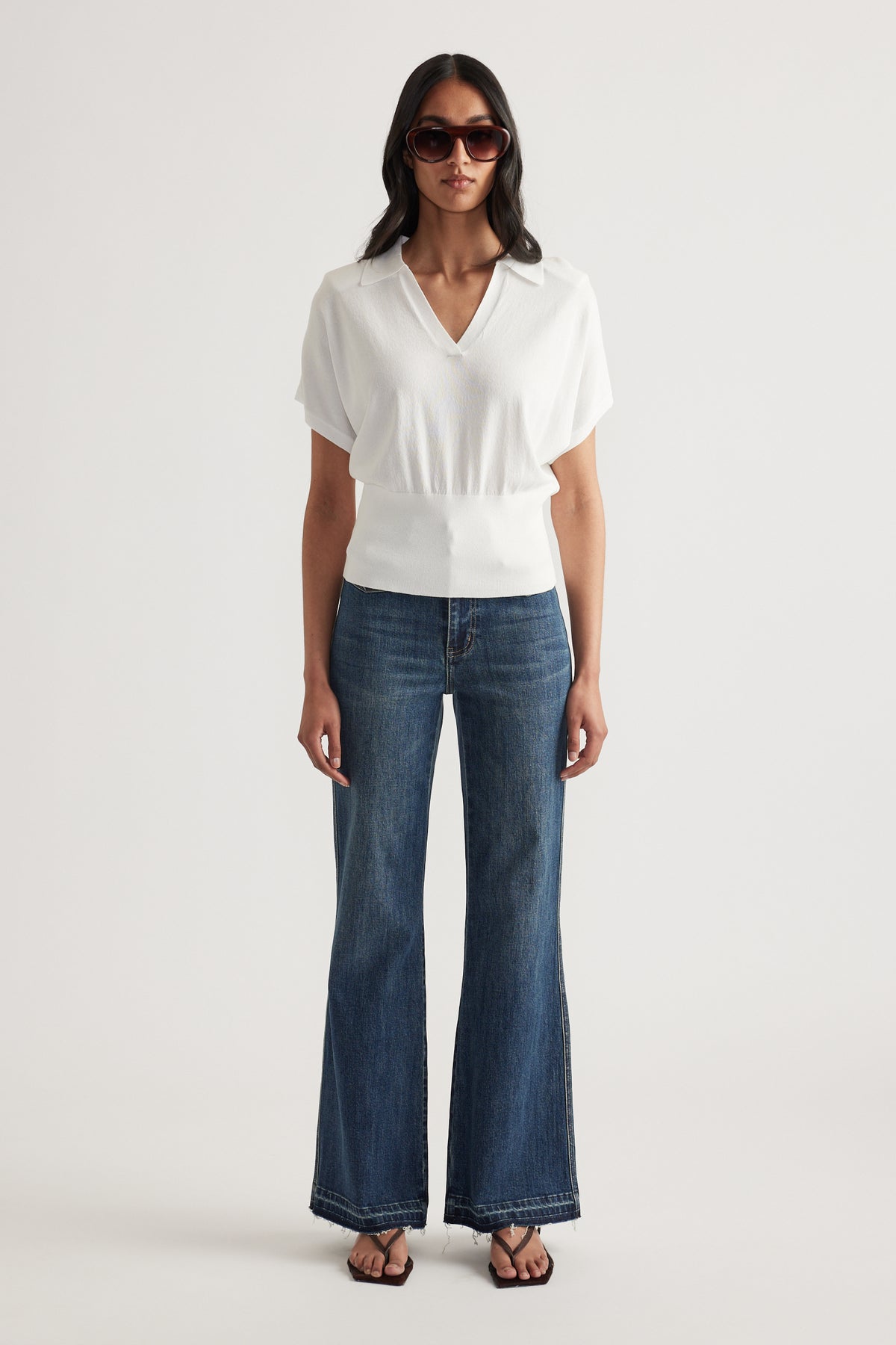 Woman wearing a white short sleeve collared knit top with a cinched waist, wide leg jeans and sunglasses