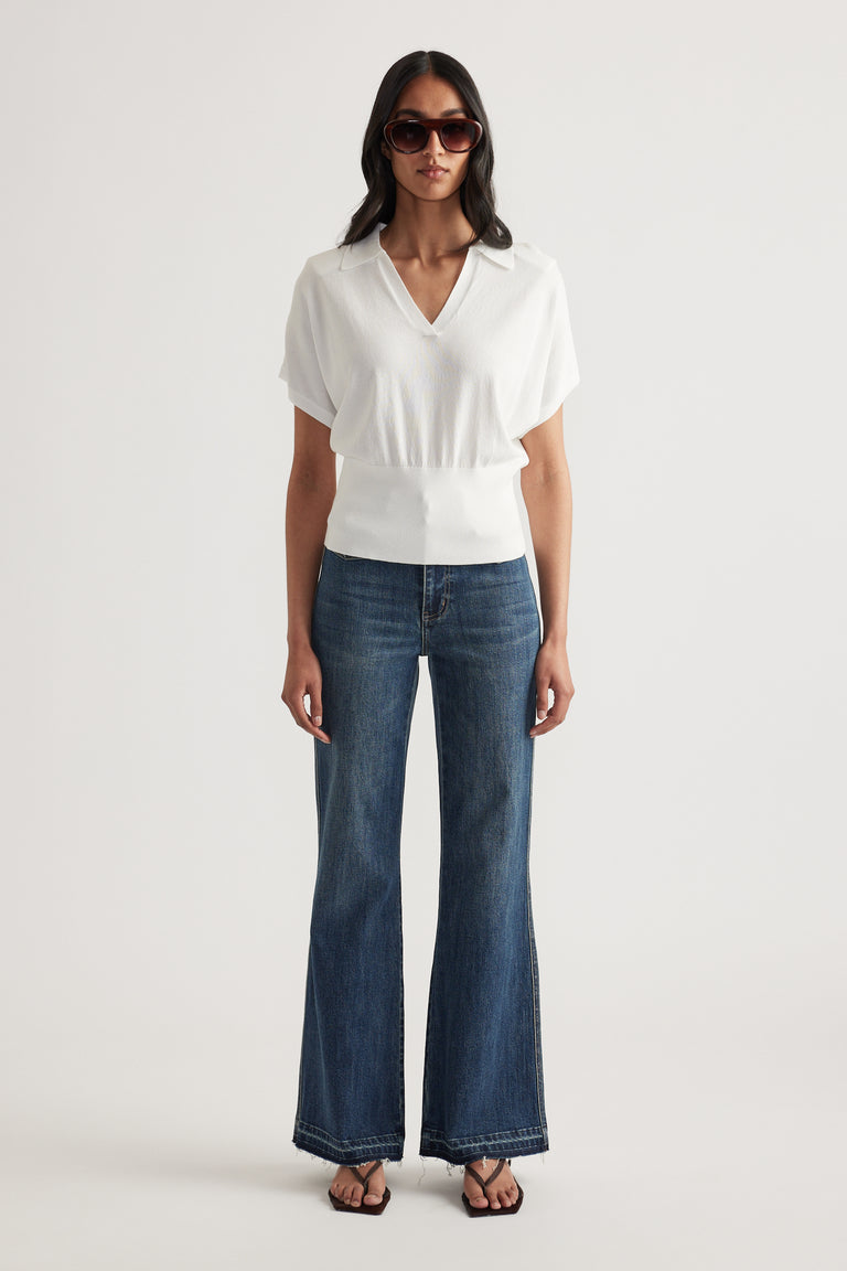 Woman wearing a white short sleeve collared knit top with a cinched waist, wide leg jeans and sunglasses