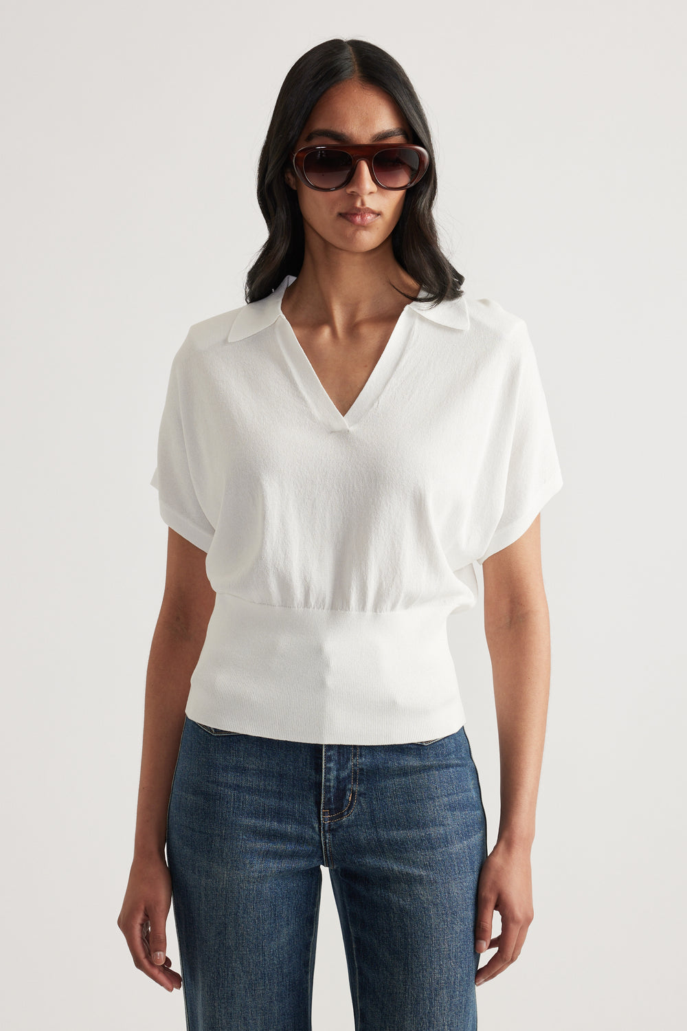 Woman wearing a white short sleeve collared V neck knit top with a cinched waist and blue jeans