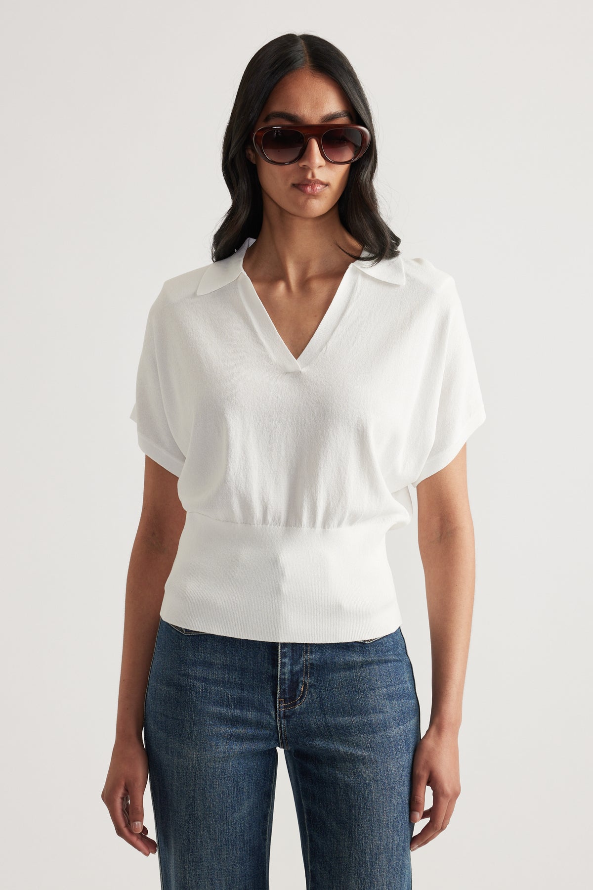 Woman wearing a white short sleeve collared V neck knit top with a cinched waist and blue jeans