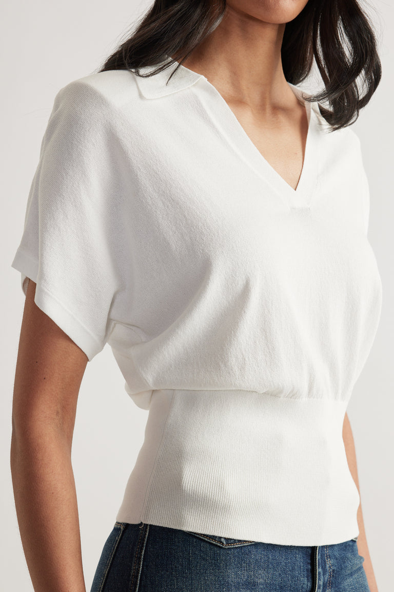 Woman wearing a white short sleeve collared V neck knit top with a cinched waist and blue jeans
