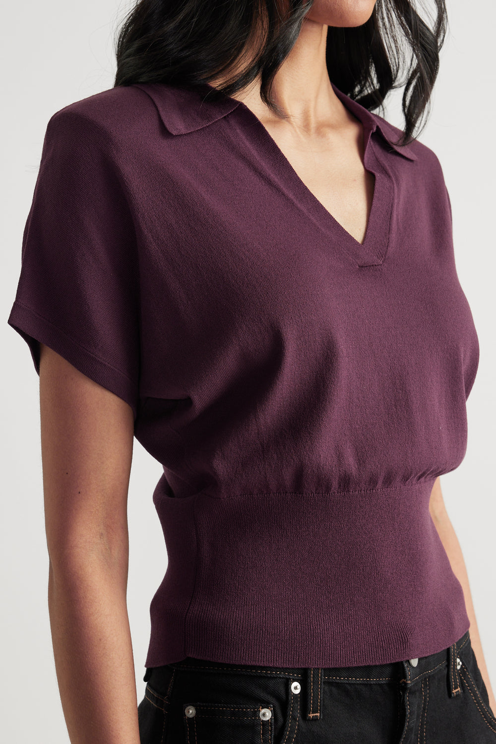 Woman wearing a burgundy short sleeve collared V neck knit top with a cinched waist and black jeans