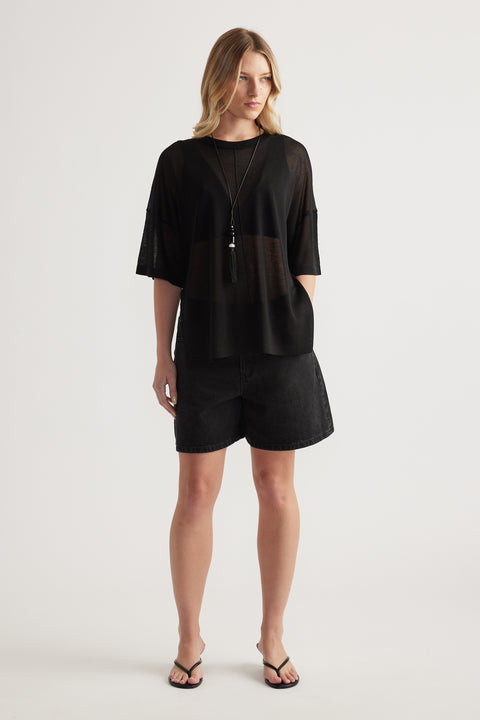Woman wearing a black sheer relaxed fit knit tee with crew neck, black shorts and flat sandals