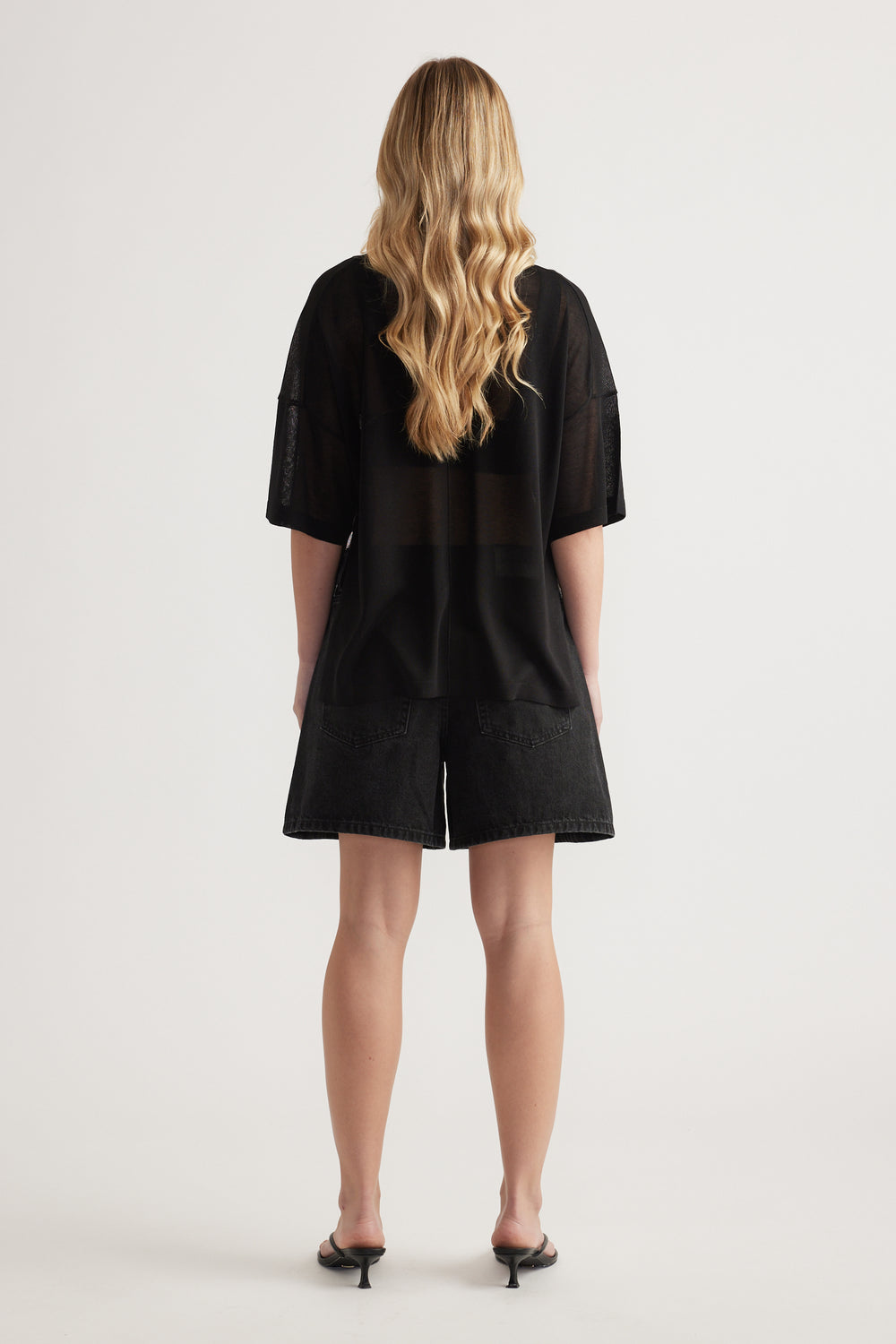 Back view of woman wearing a relaxed black longline knit tee with short sleeves and sheer detailing