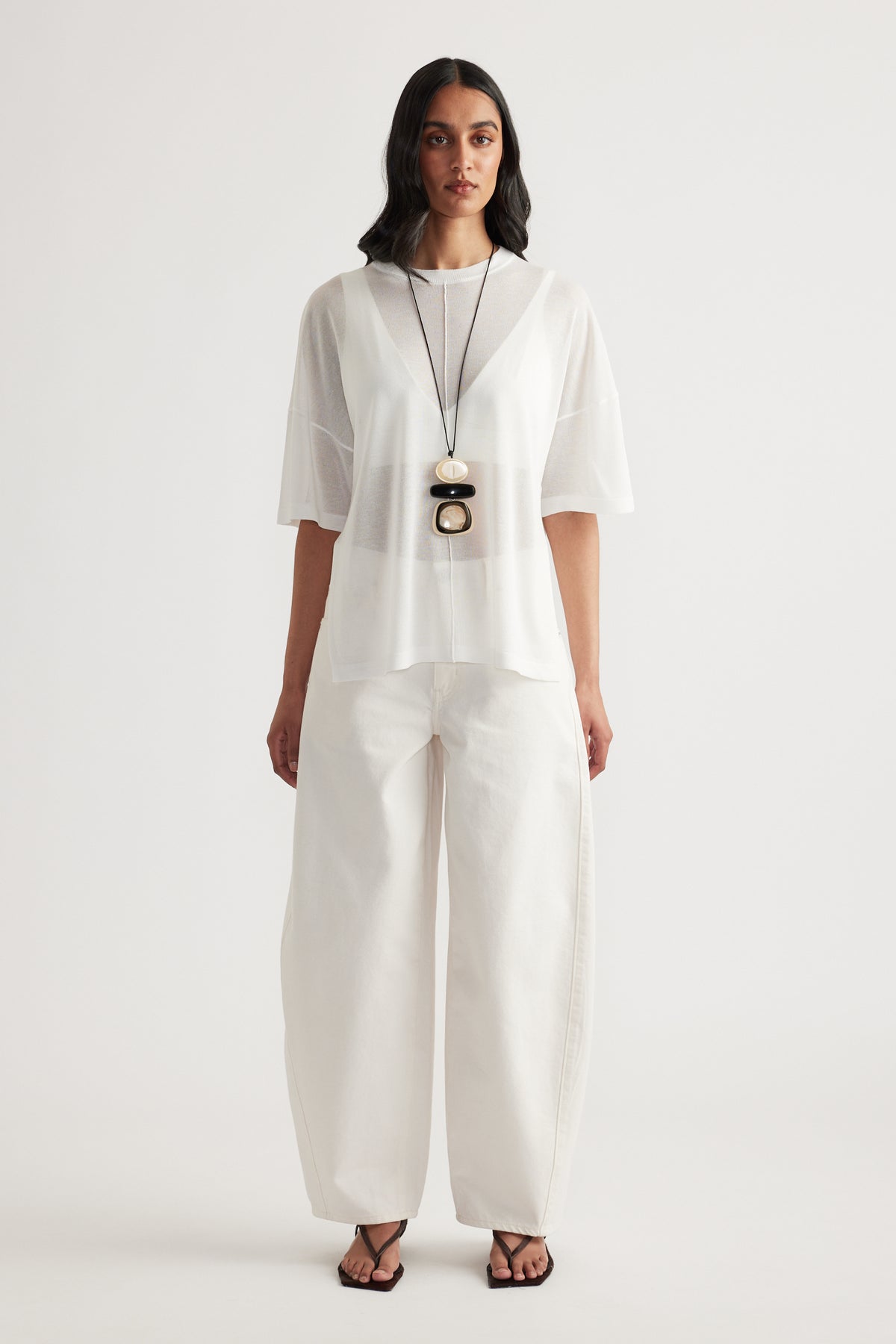 Woman wearing a sheer white oversized knit tee with wide leg white pants and layered pendant necklace