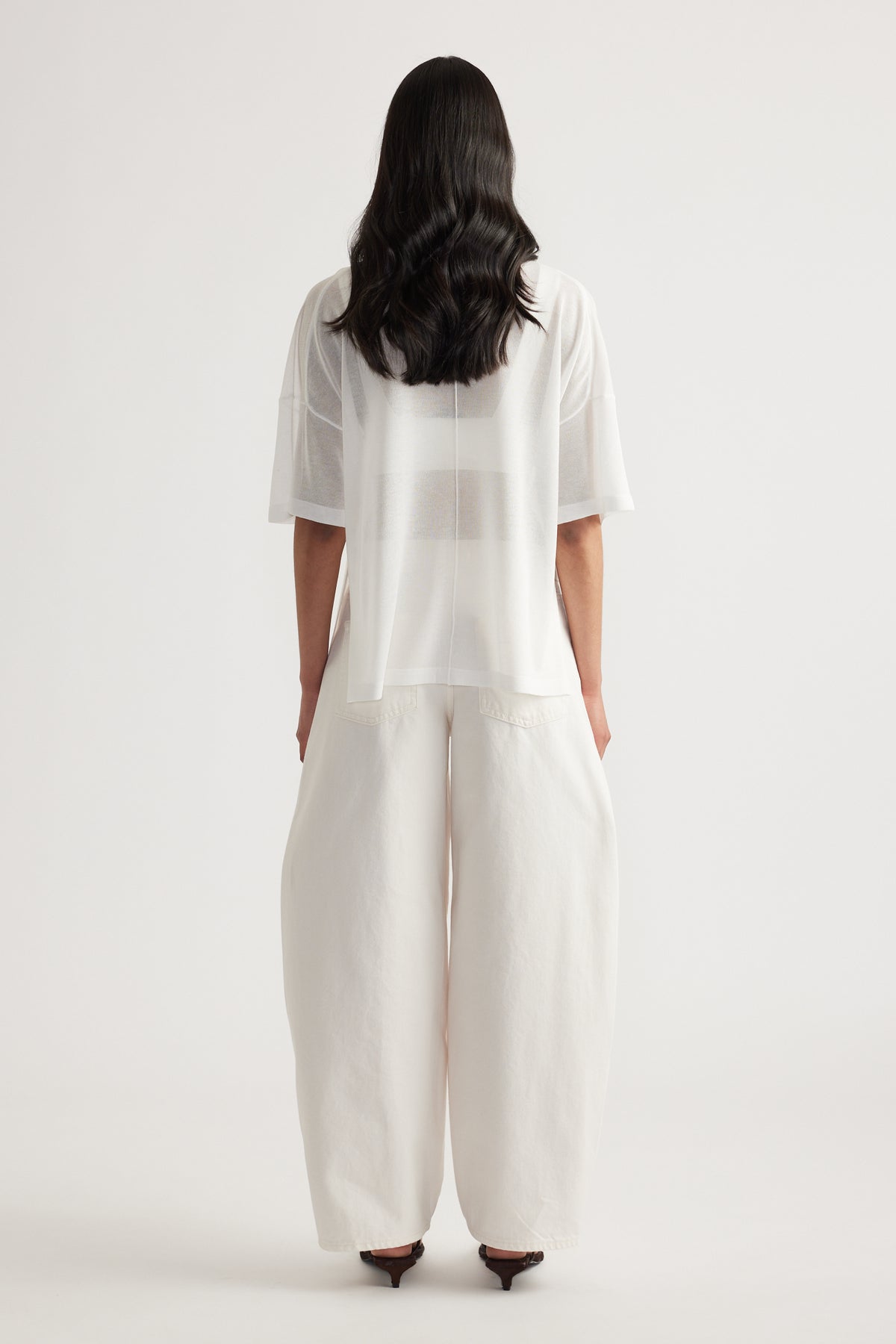 Back view of woman wearing a sheer white relaxed fit longline knit tee with short sleeves and wide leg pants