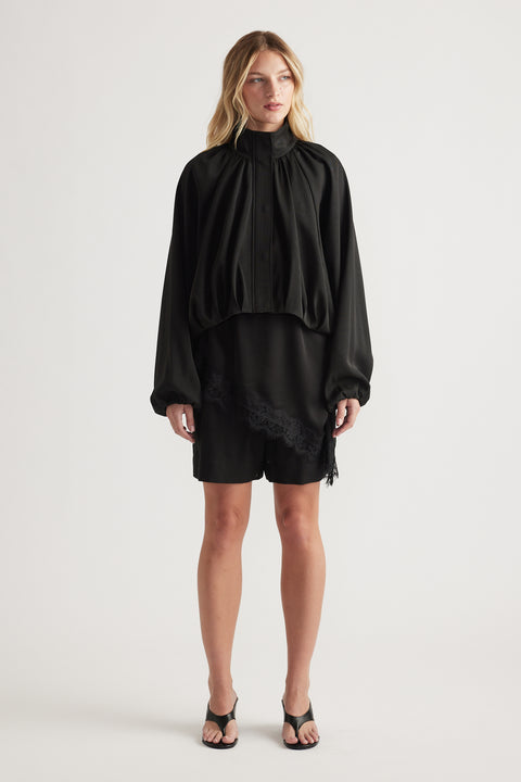 Woman wearing a loose black blouse with voluminous sleeves and black shorts featuring subtle lace trim, paired with black heeled sandals.