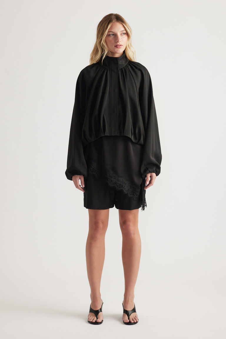 Woman wearing a loose black blouse with voluminous sleeves and black shorts featuring subtle lace trim, paired with black heeled sandals.