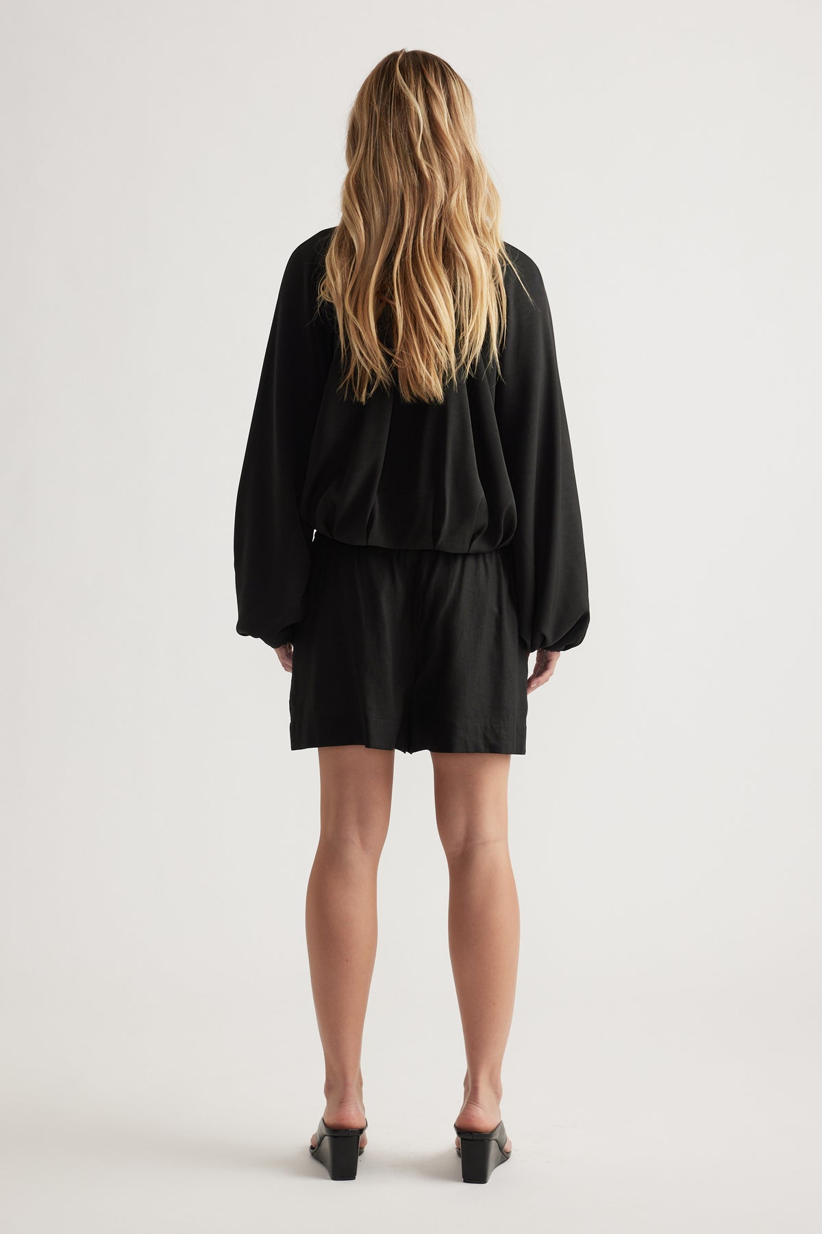 Woman with loose wavy hair wearing black linen blend shorts paired with a loose long-sleeve top and heeled sandals viewed from behind