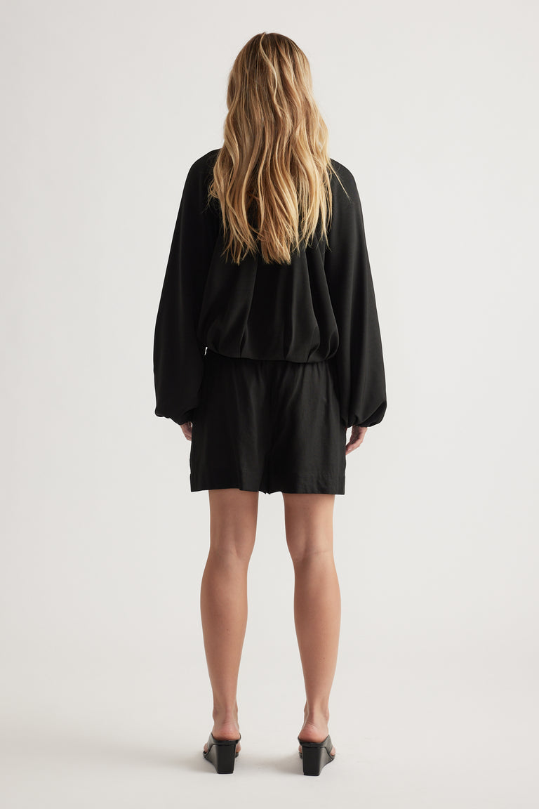 Woman with loose wavy hair wearing black linen blend shorts paired with a loose long-sleeve top and heeled sandals viewed from behind