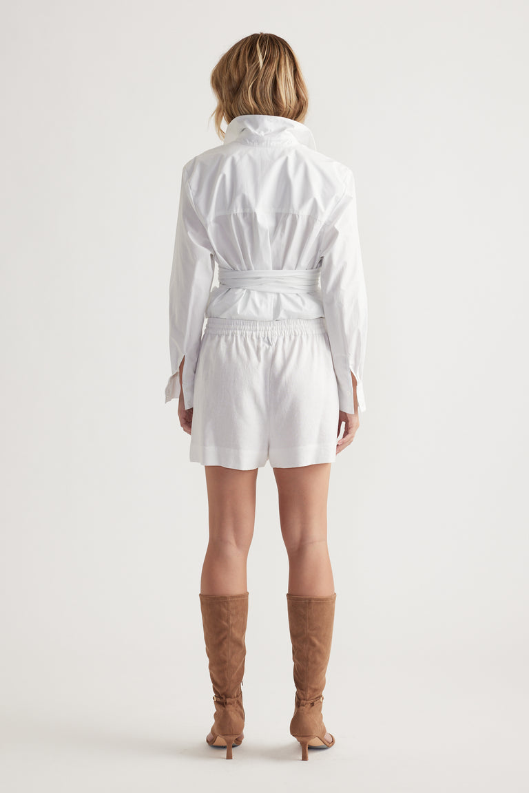 Woman seen from behind wearing white tailored shorts paired with a cinched waist shirt and tan knee-high boots