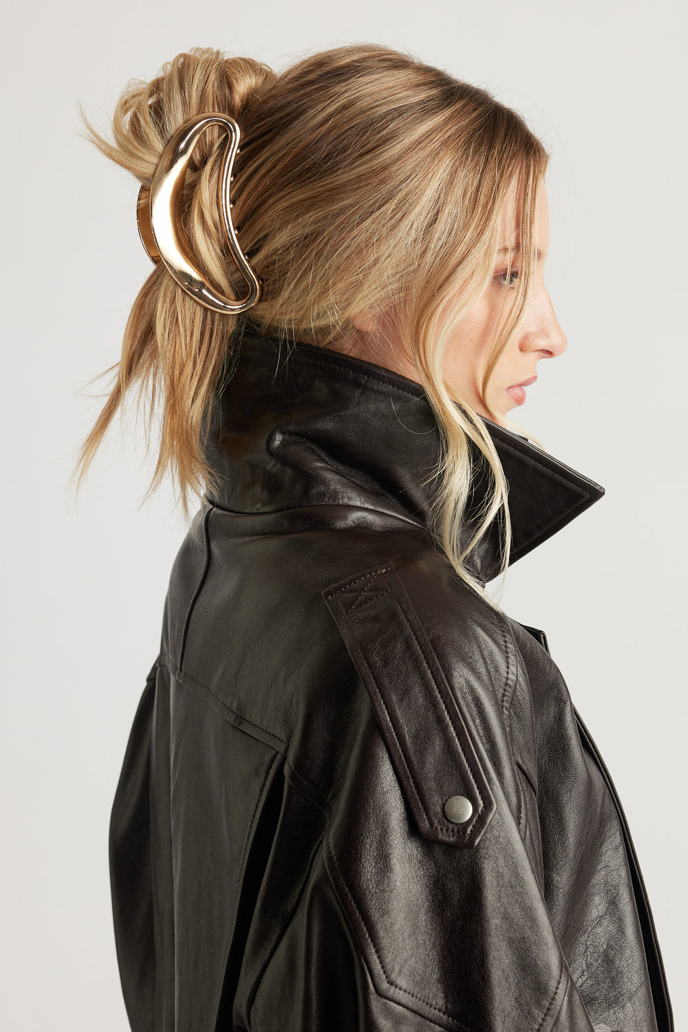 Profile view of a woman with blonde hair secured by a gold-toned hair claw, wearing a high-collar black leather jacket