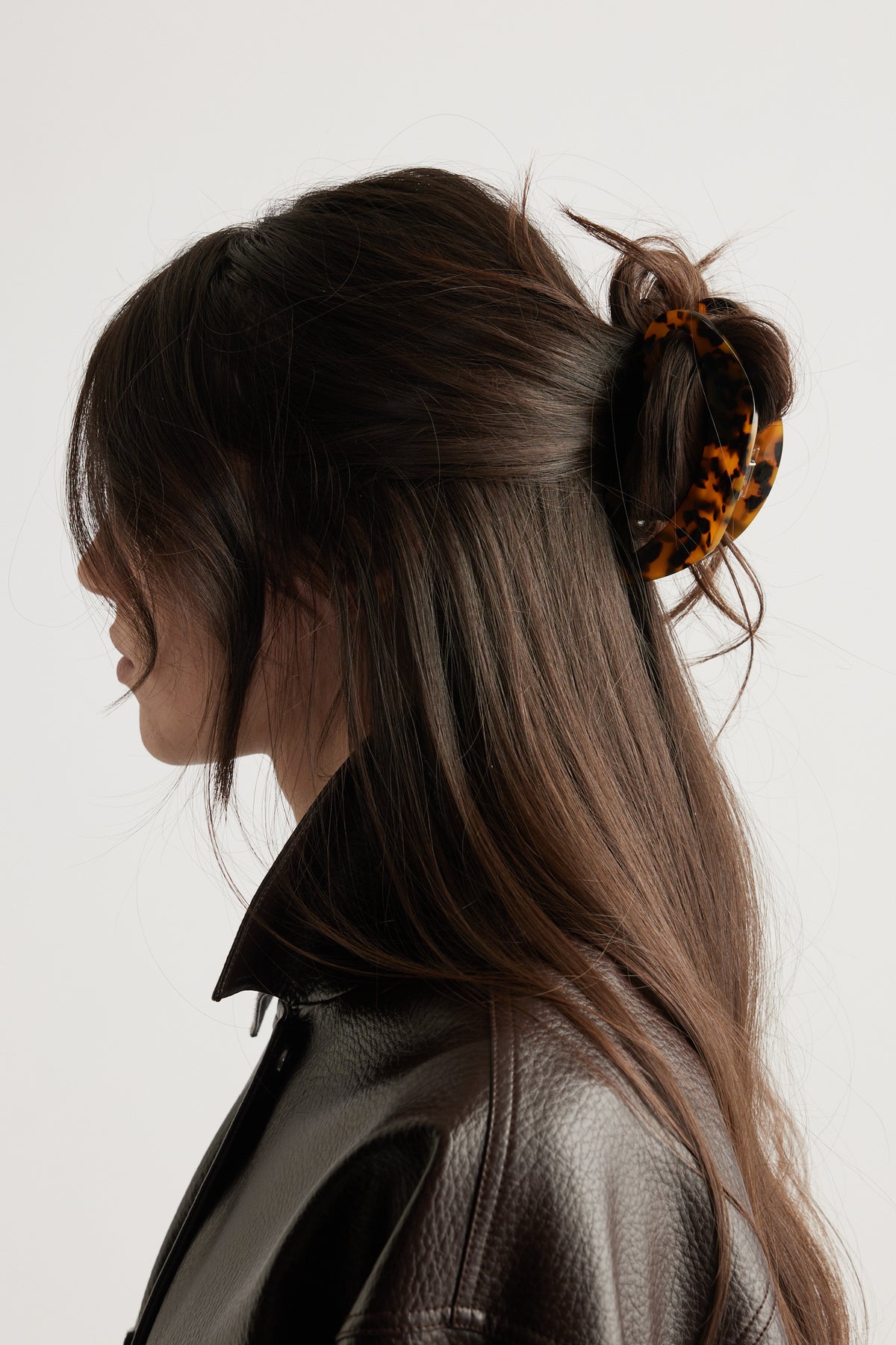 Side profile of a woman with long dark hair styled half up with a tortoiseshell hair claw and wearing a brown leather jacket