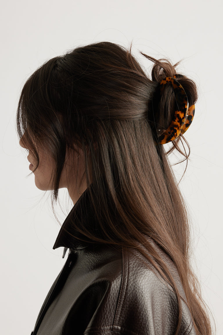 Side profile of a woman with long dark hair styled half up with a tortoiseshell hair claw and wearing a brown leather jacket