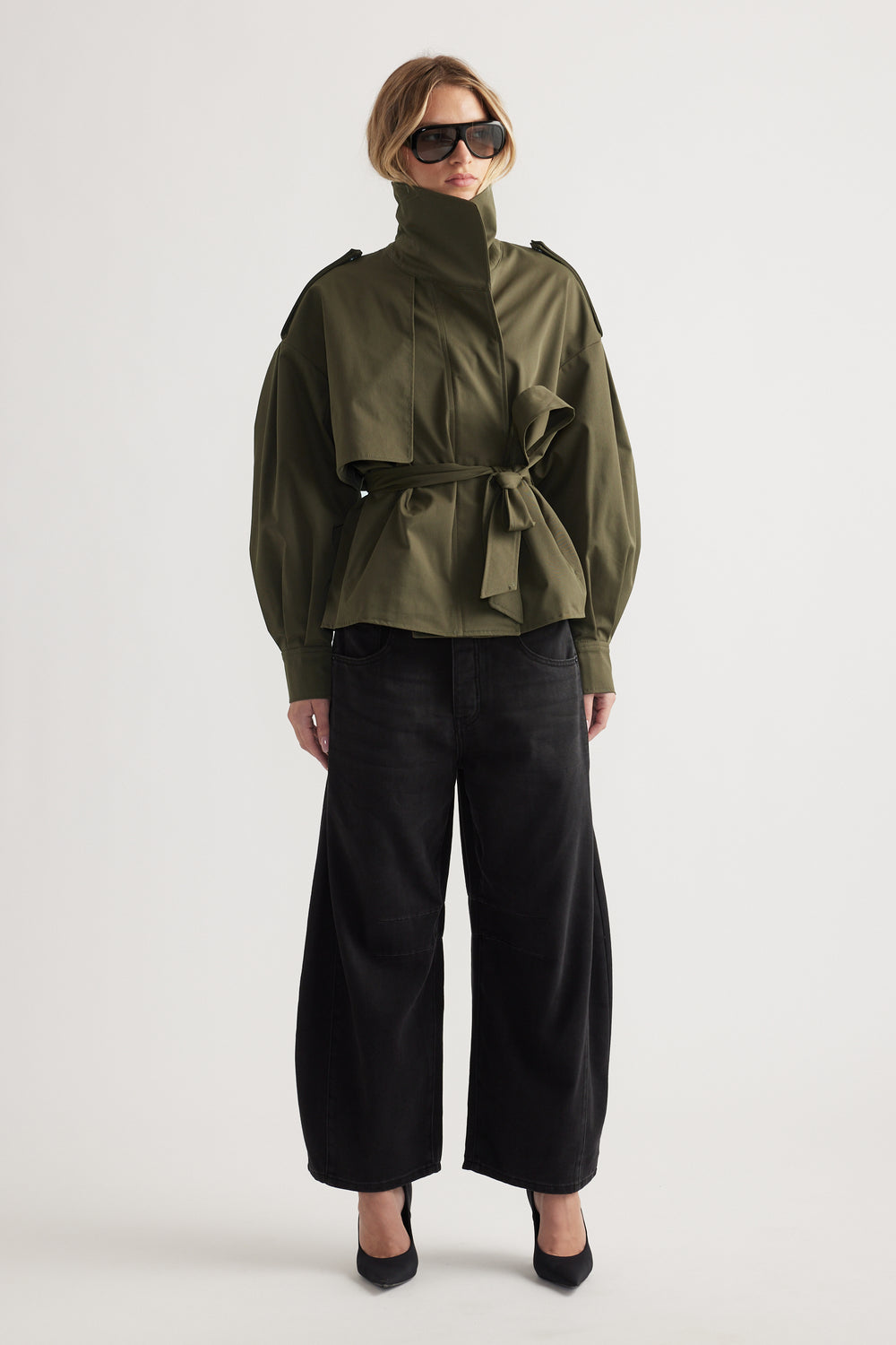 Woman wearing high-waisted wide-leg black jeans paired with an olive belted jacket and oversized sunglasses