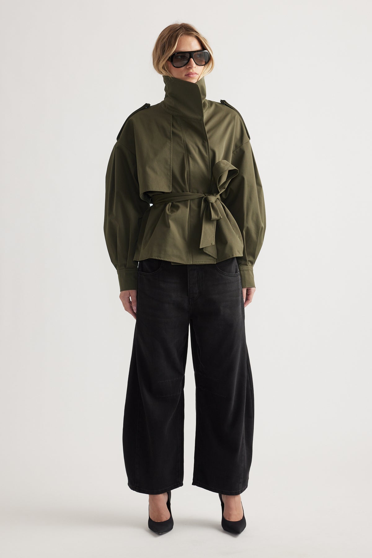Woman wearing high-waisted wide-leg black jeans paired with an olive belted jacket and oversized sunglasses