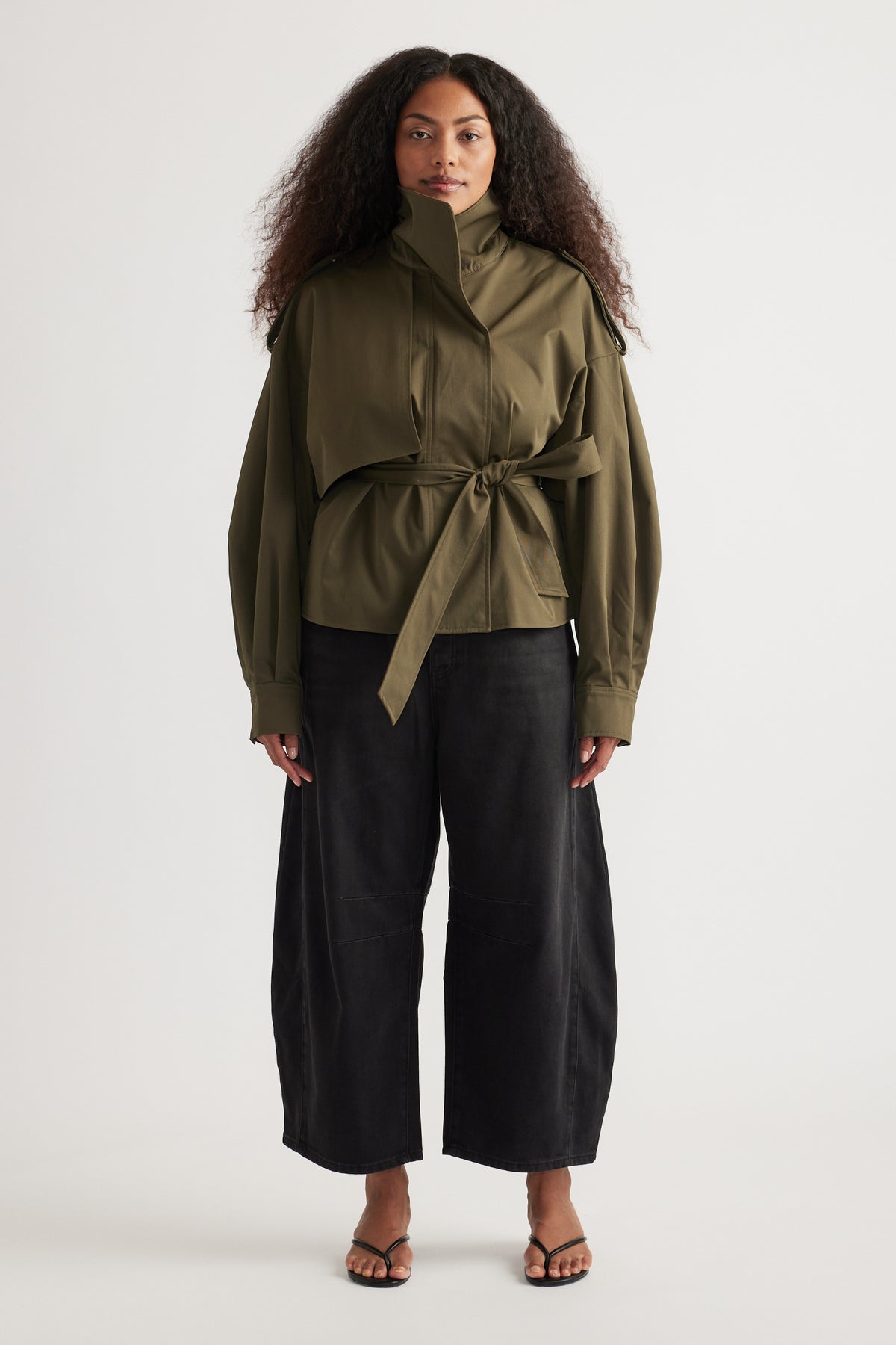 Woman wearing an olive belted jacket with black cropped barrel-leg jeans and flat sandals