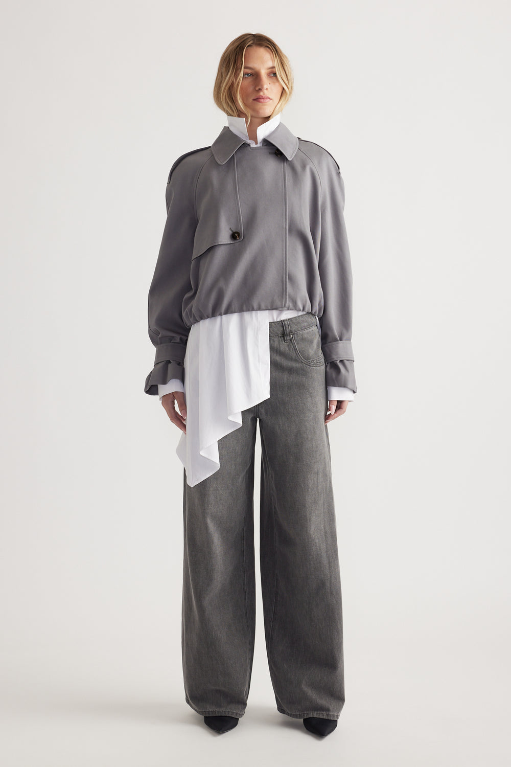 Woman wearing a grey cropped jacket over a white asymmetrical blouse paired with wide-leg grey denim jeans and black shoes