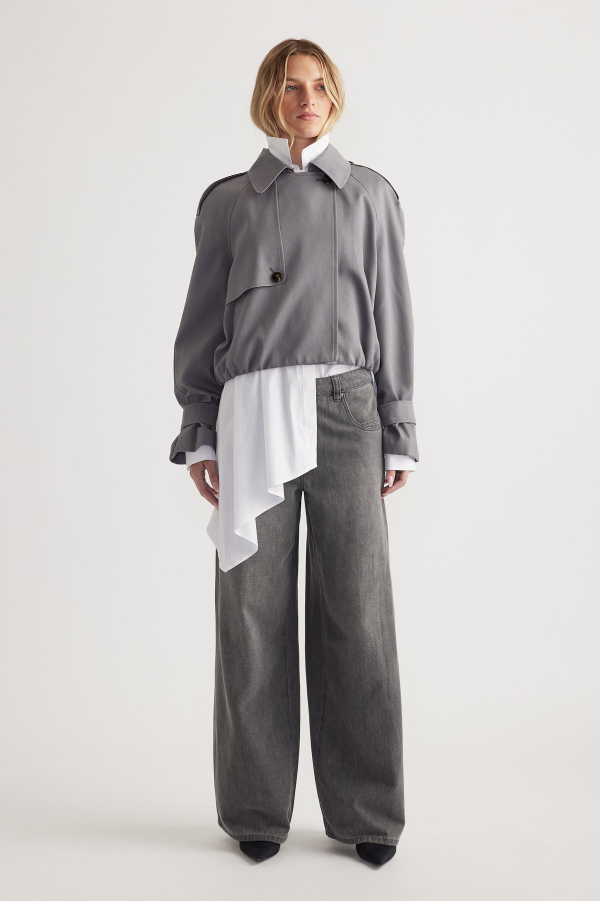 Woman wearing a grey cropped jacket over a white asymmetrical blouse paired with wide-leg grey denim jeans and black shoes