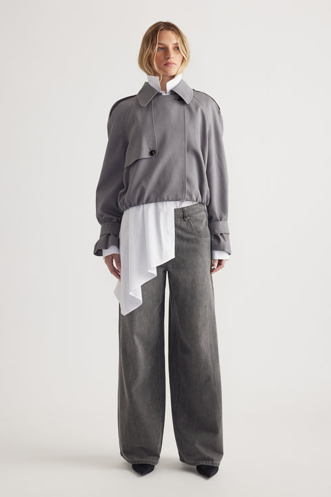 Woman wearing a grey cropped jacket over a white asymmetrical blouse paired with wide-leg grey denim jeans and black shoes