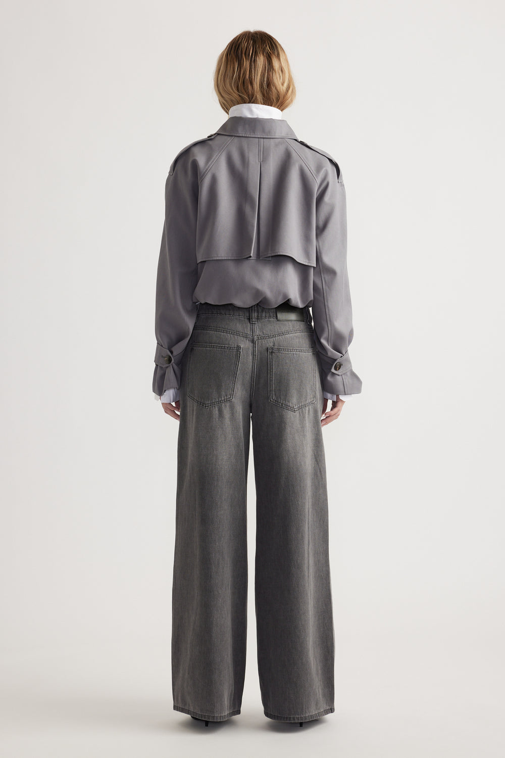 Woman wearing grey cropped jacket paired with mid-rise wide leg denim jeans, seen from behind on plain background