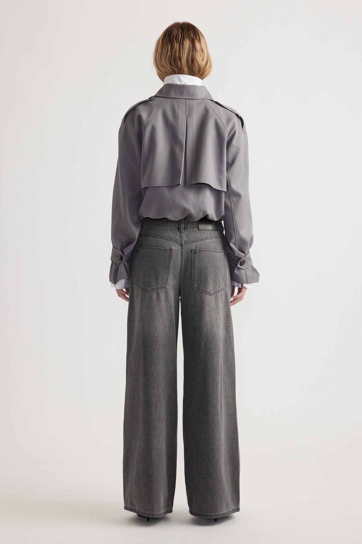 Woman wearing grey cropped jacket paired with mid-rise wide leg denim jeans, seen from behind on plain background