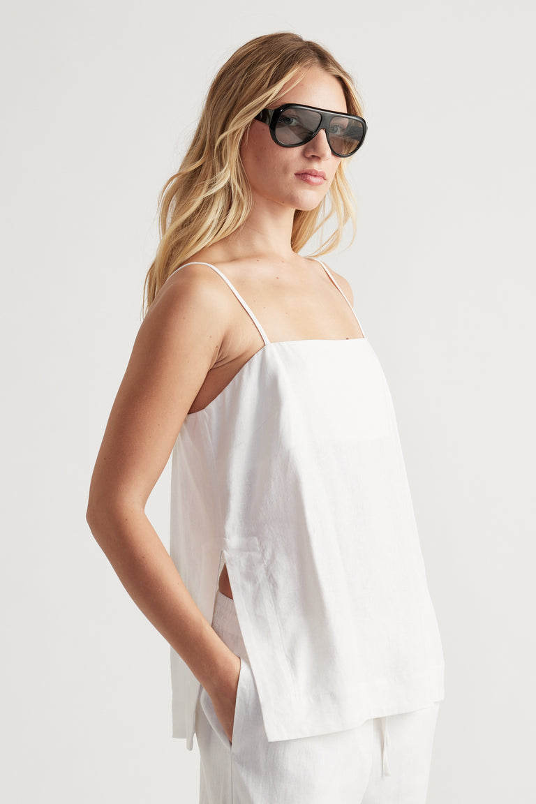 Woman wearing a white linen blend cami with thin straps and side slits paired with matching relaxed pants and sunglasses
