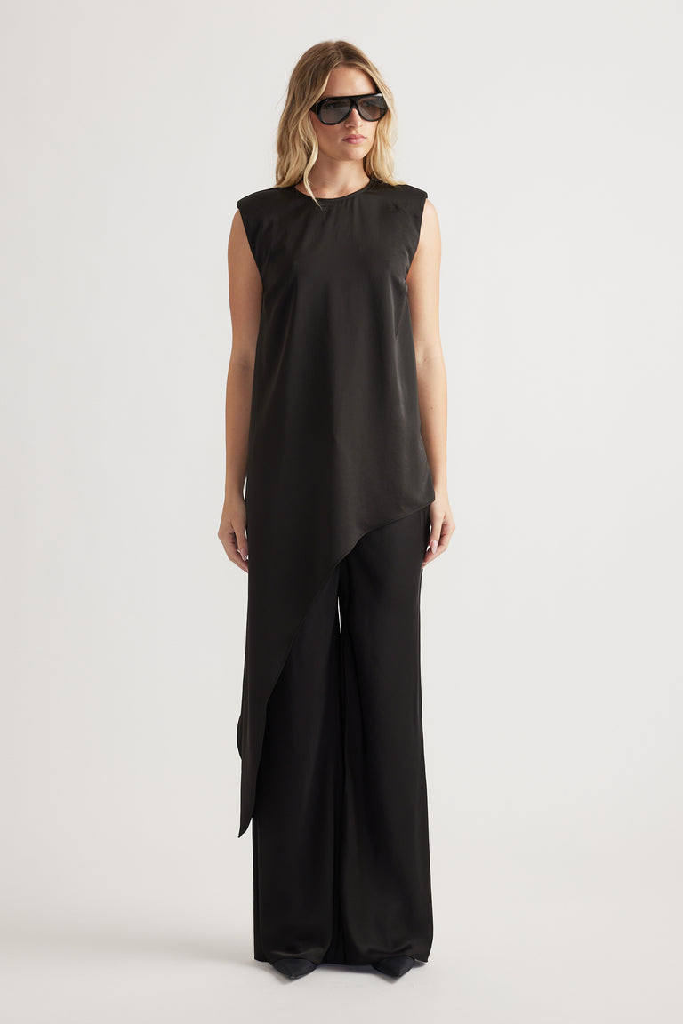 Woman wearing a sleeveless black drape top with asymmetric hem paired with wide-leg black pants and oversized sunglasses