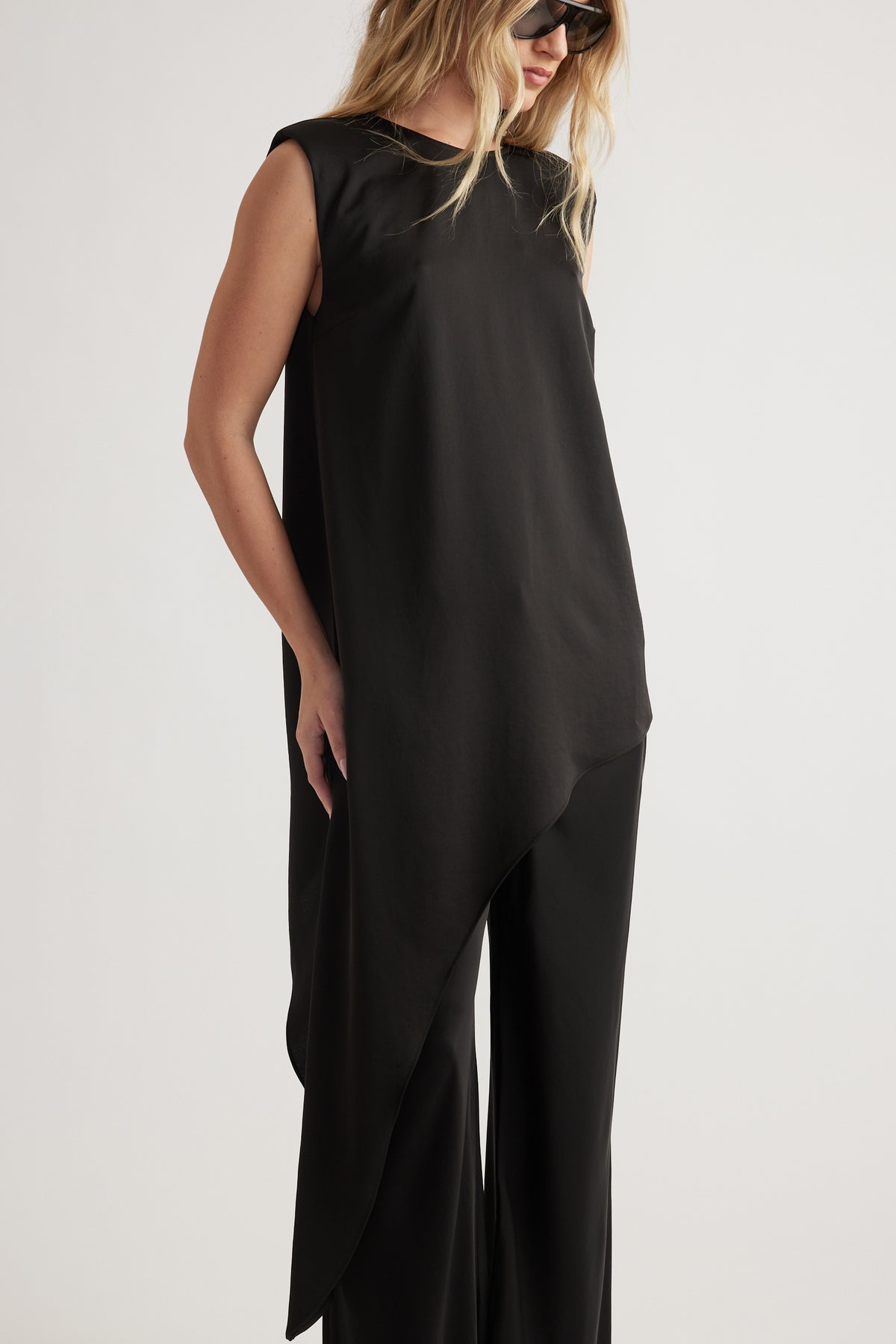 Woman wearing an asymmetrical drape black top paired with wide-leg black pants and sunglasses in a minimalist style