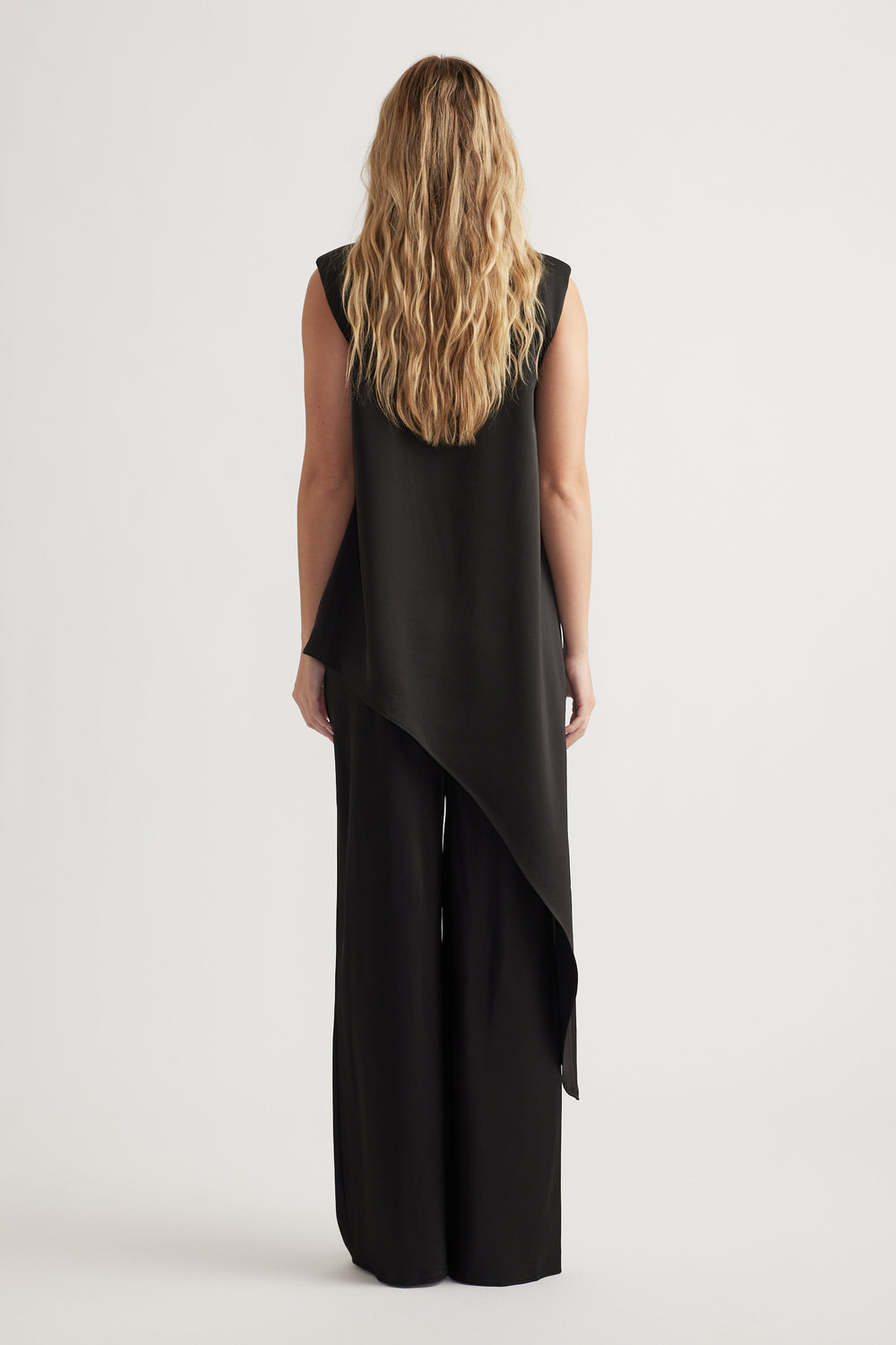 Woman seen from behind wearing a sleeveless black draped top with asymmetrical hem paired with wide-leg black pants
