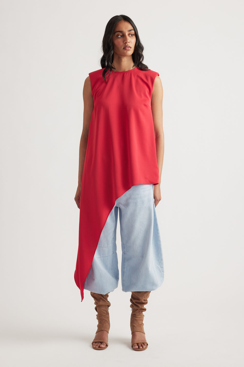 Woman wearing a sleeveless crimson drape top with an asymmetrical hem, paired with light wide-leg pants and tan boots