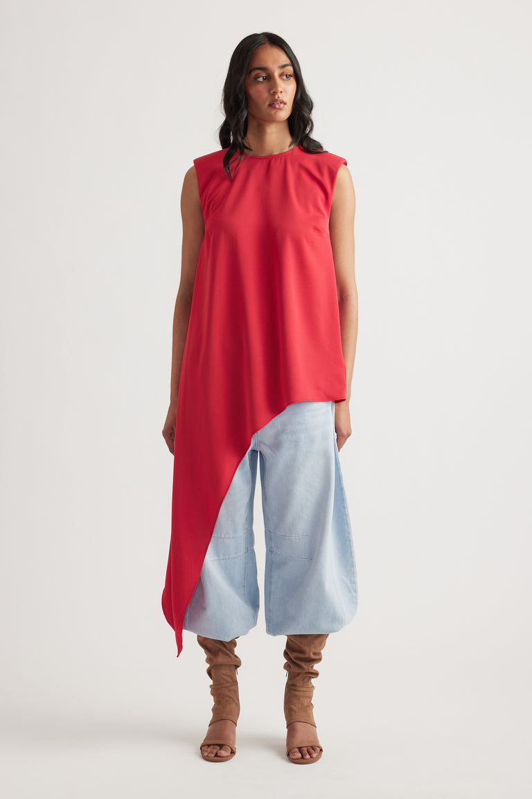 Woman wearing a sleeveless crimson drape top with an asymmetrical hem, paired with light wide-leg pants and tan boots