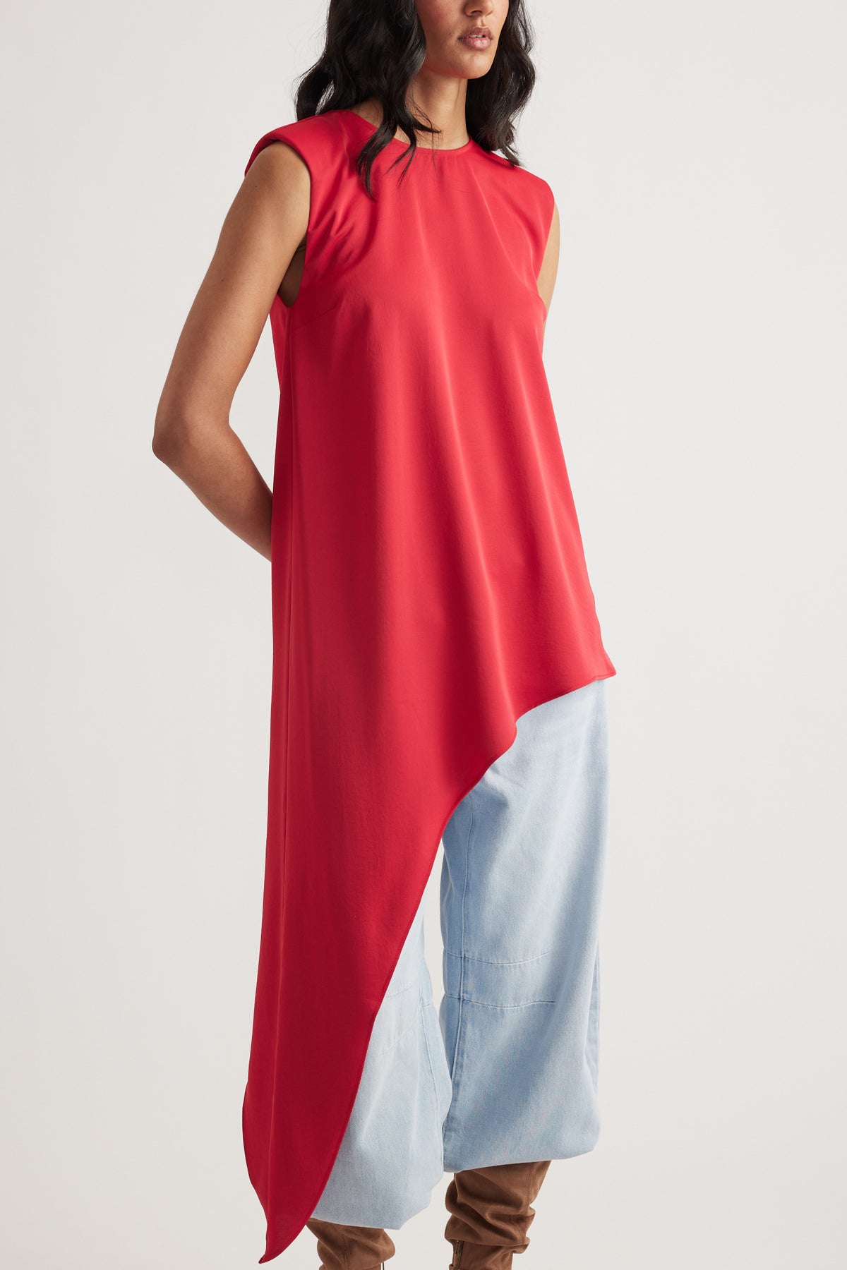 Woman wearing a sleeveless red drape top with asymmetrical hem paired with light blue wide-leg pants and brown boots