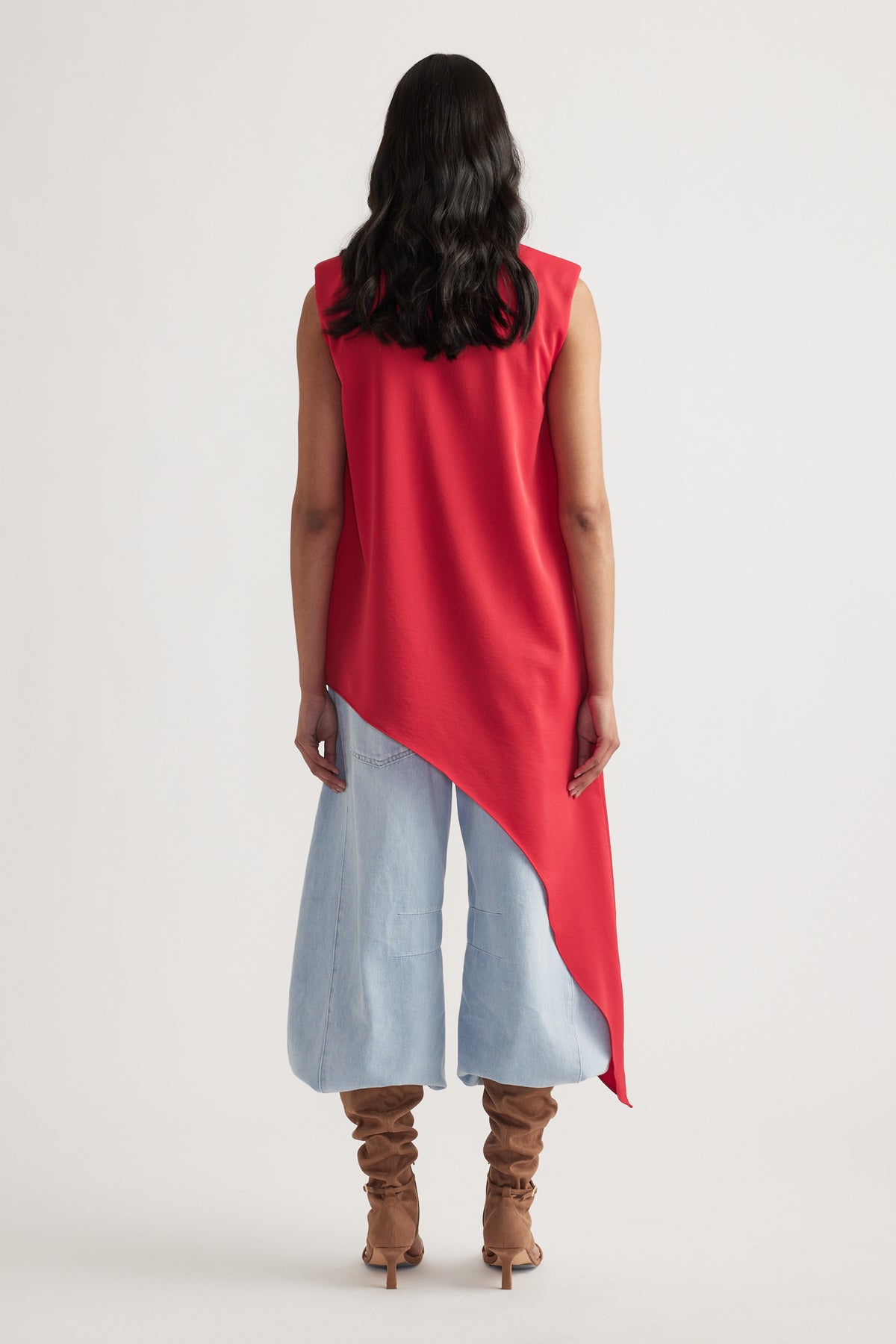 Woman seen from behind wearing an asymmetrical crimson sleeveless top paired with light wide-leg jeans and slouched tan boots