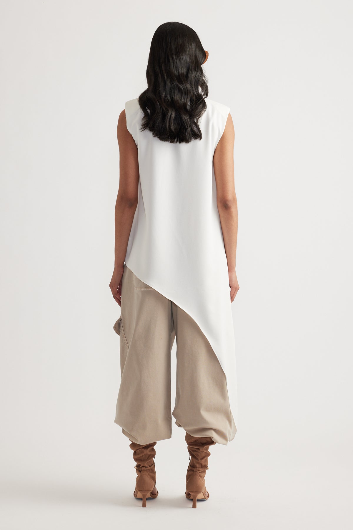 Woman seen from behind wearing an ivory sleeveless drape top paired with loose beige pants and tan heeled boots