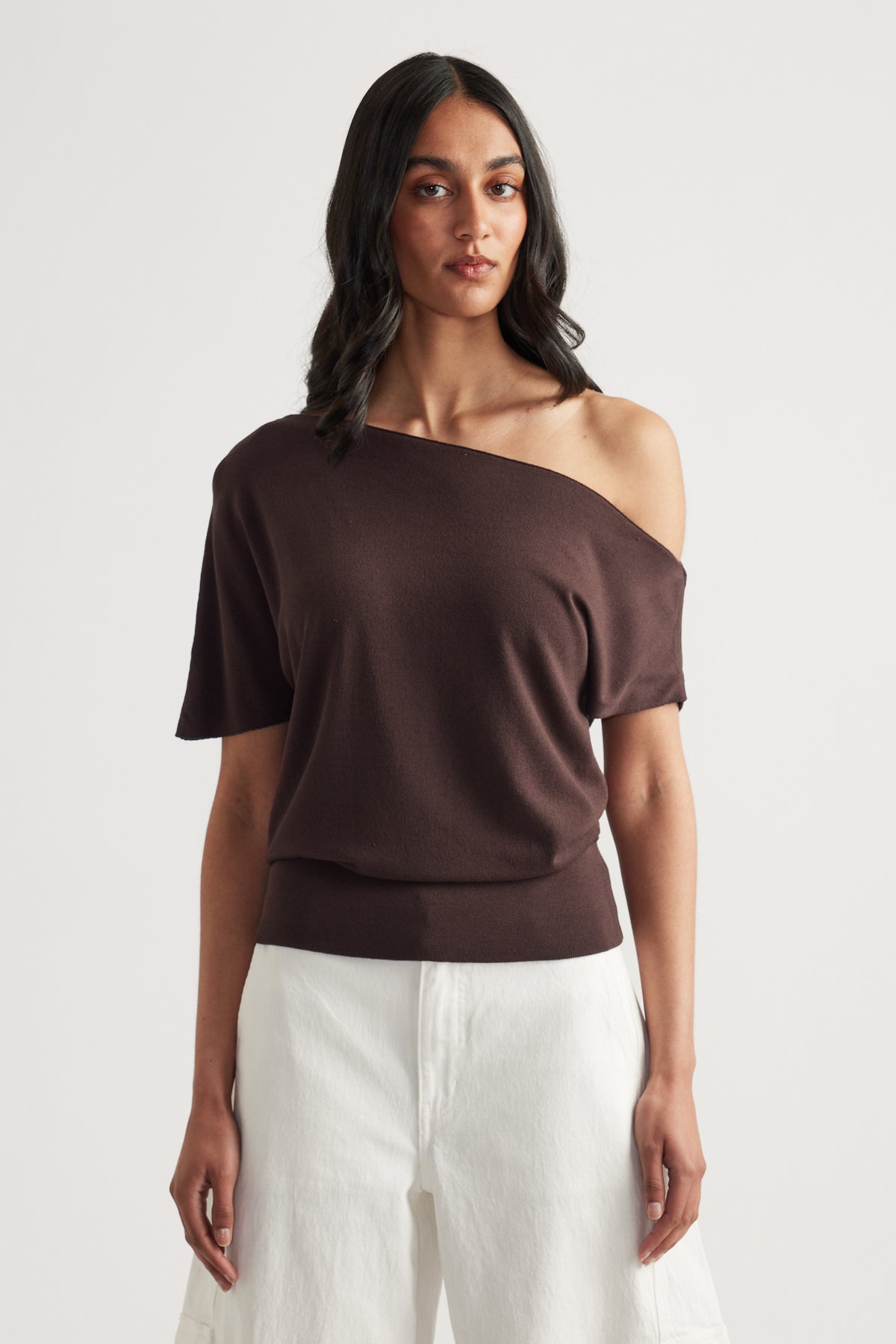 Woman wearing a brown asymmetric off shoulder knit top with cinched hem and white pants