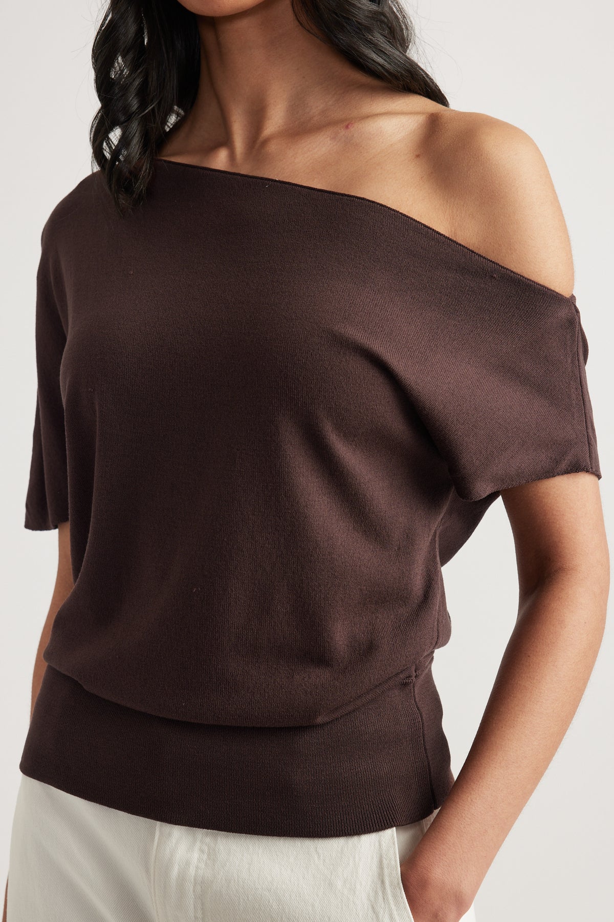 Woman wearing a dark brown off shoulder knit top with asymmetric neckline and cinched hem, paired with cream pants