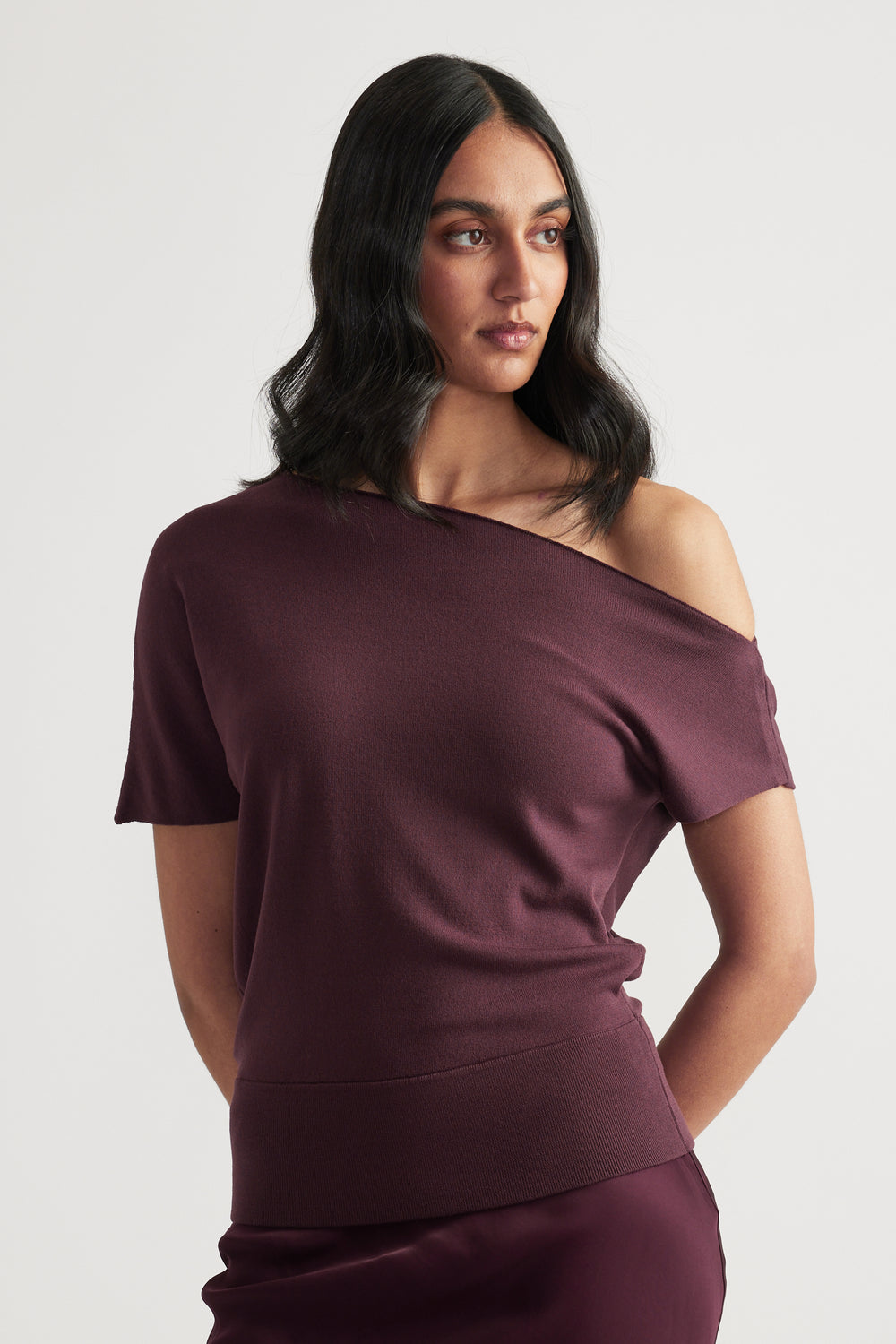 Model wearing a burgundy lightweight knit top with an asymmetric off shoulder neckline and cinched hem