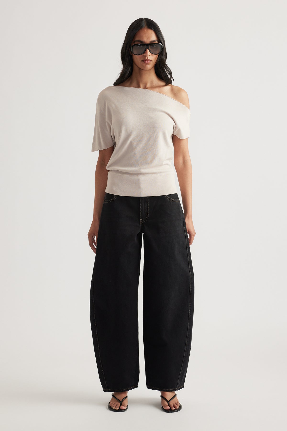 Woman wearing a light beige off shoulder knit top with cinched hem, black wide leg pants, sunglasses and sandals
