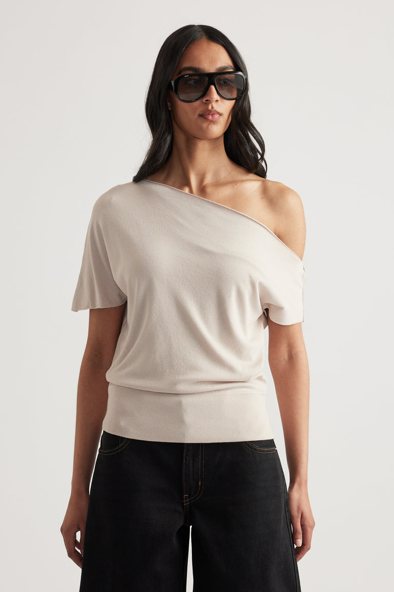 Woman wearing a cream off shoulder knit top with asymmetric neckline and cinched hem, paired with black jeans