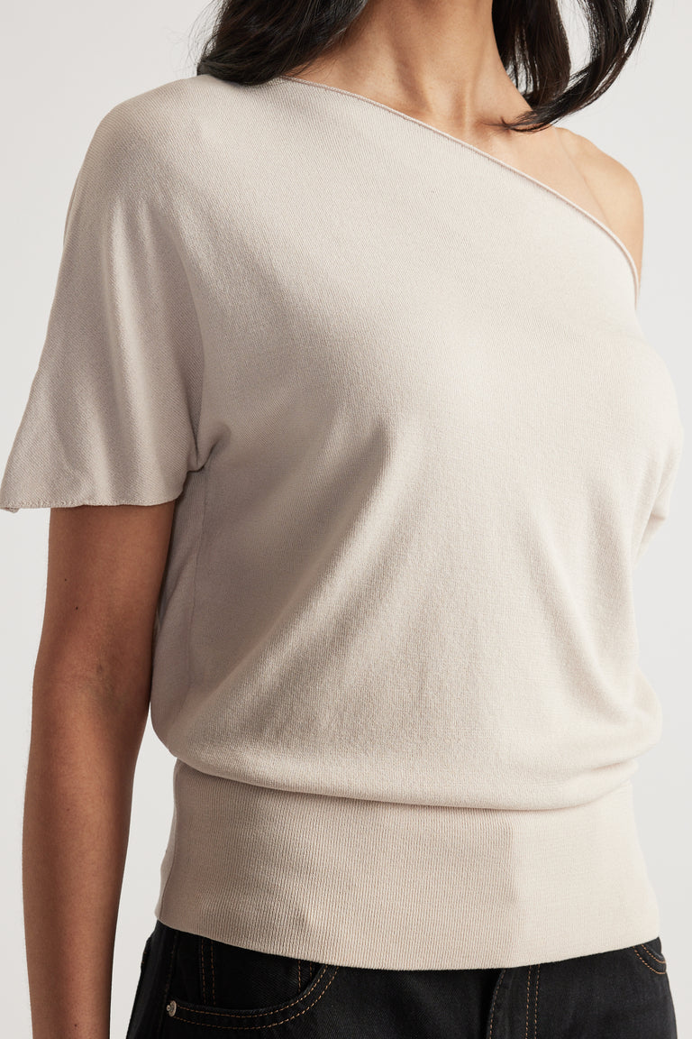 Woman wearing a cream knit top with an asymmetric off shoulder neckline short sleeves and cinched hem