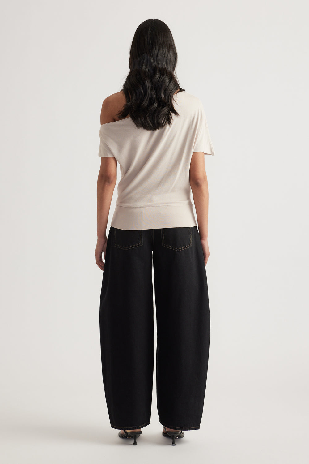 Woman wearing an asymmetric off shoulder cream knit top with cinched hem and black wide leg pants viewed from behind