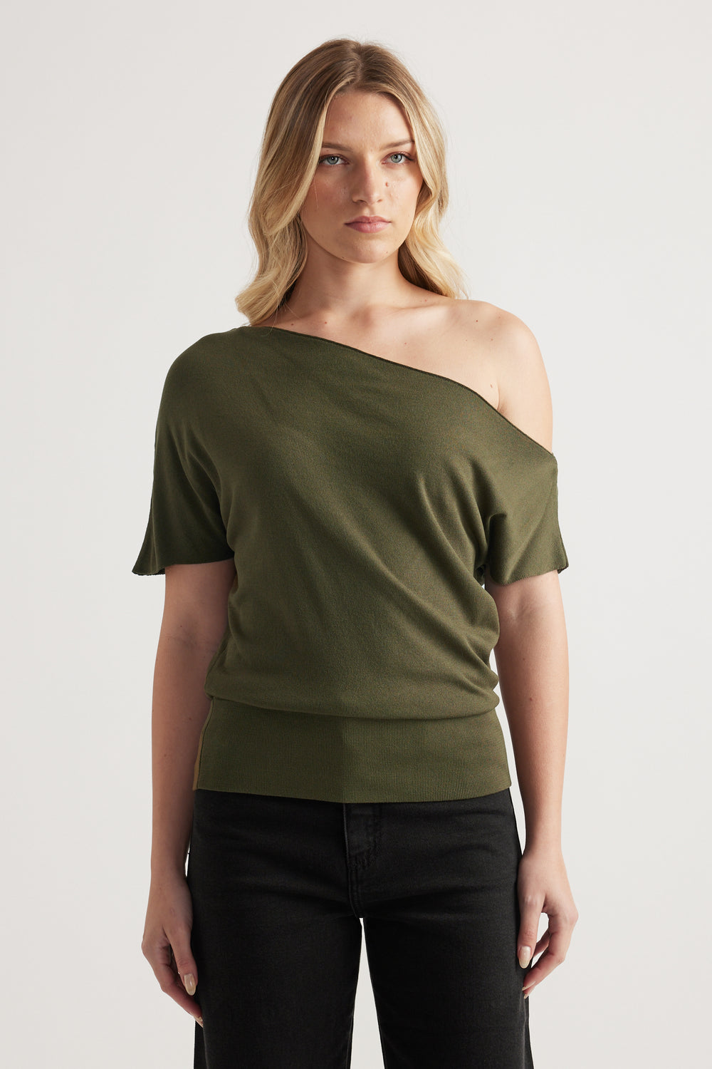 Woman wearing an olive green off shoulder knit top with asymmetric neckline and cinched hem, styled with black jeans