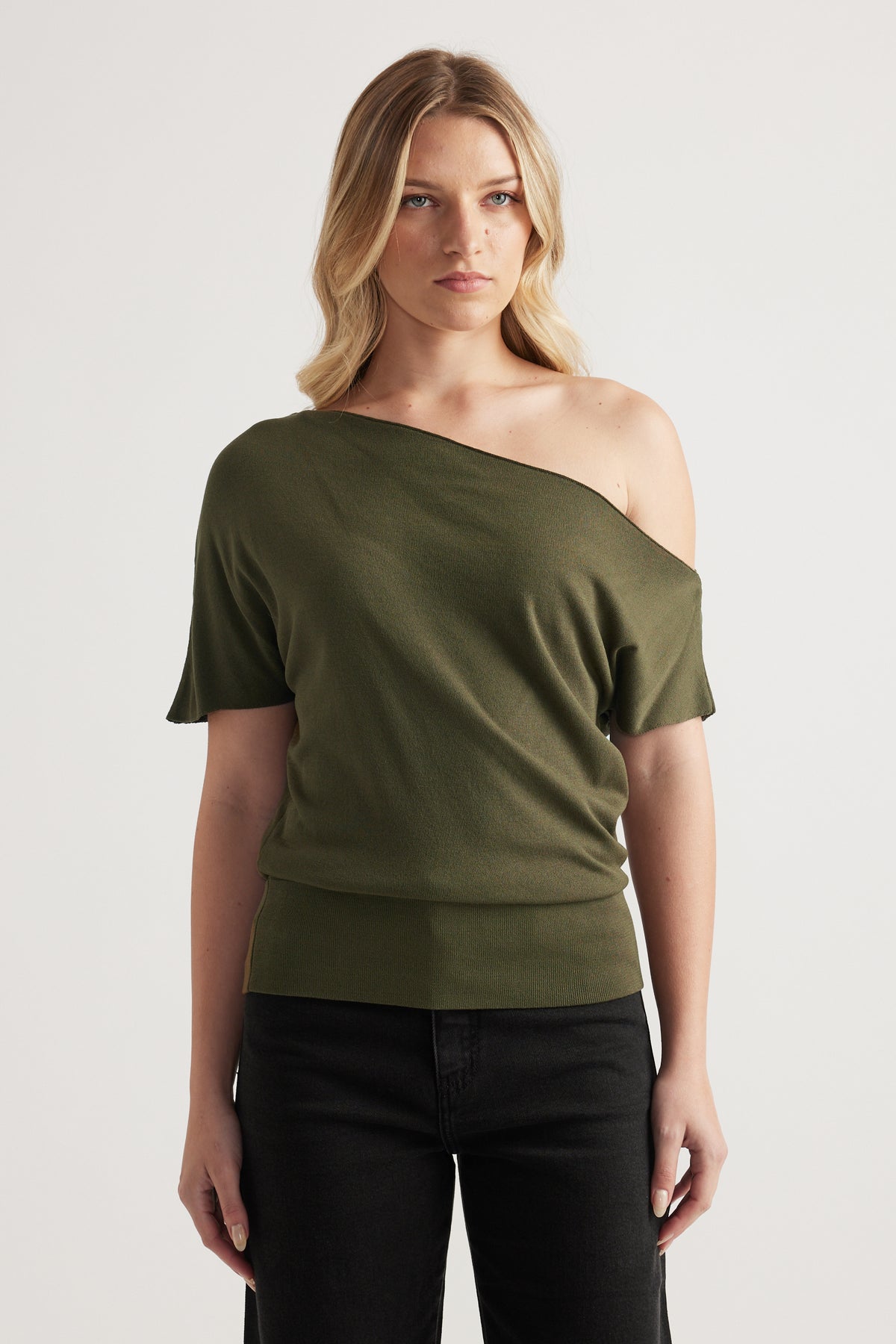 Woman wearing an olive green off shoulder knit top with asymmetric neckline and cinched hem, styled with black jeans