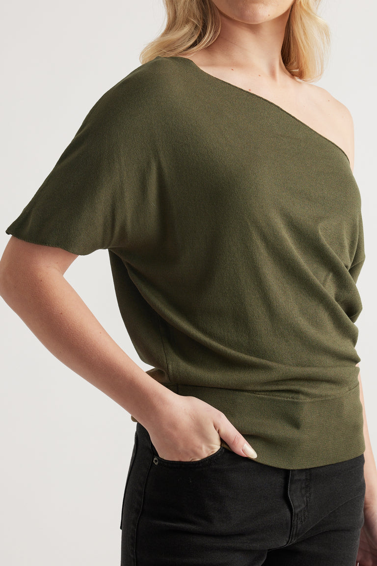 Woman wearing an olive green off shoulder knit top with asymmetric neckline and cinched hem, paired with black jeans