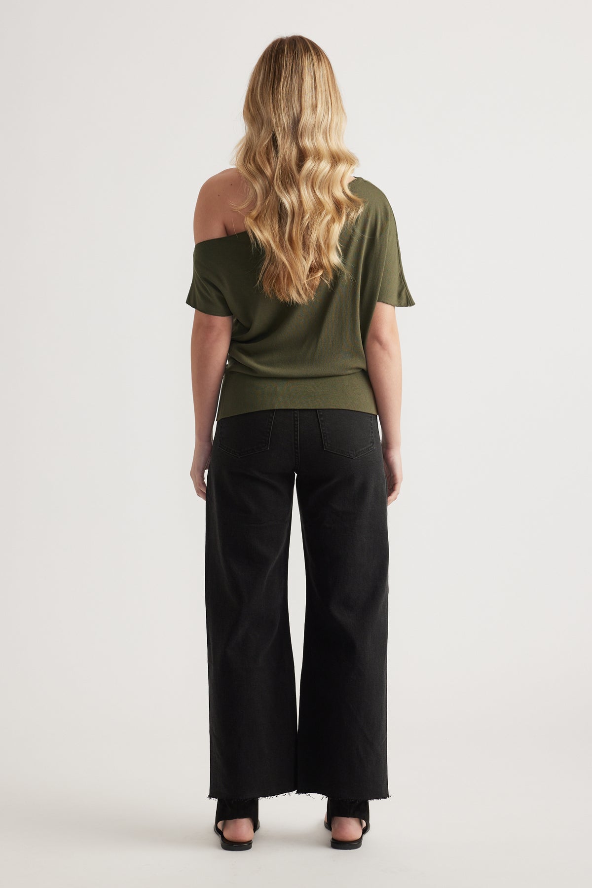 Back view of woman in an olive asymmetric off shoulder knit top with black wide leg pants and black sandals