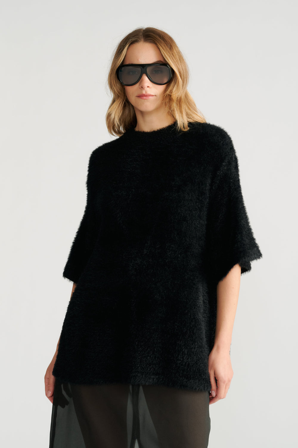 Woman wearing an oversized black knit tee with half sleeves and large black sunglasses against a plain background
