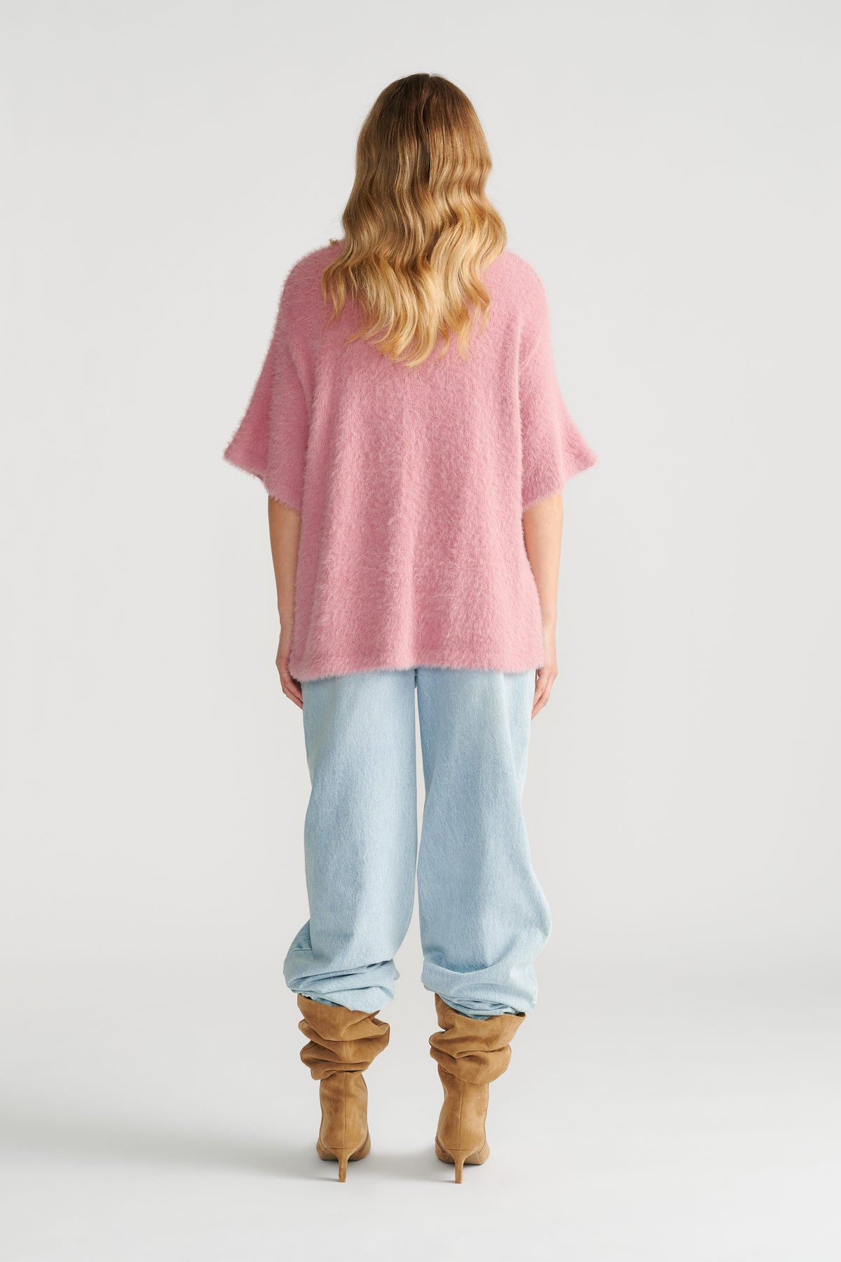 Woman wearing a soft pink oversized knit tee paired with loose light wash jeans and tan slouchy heeled boots, back view.