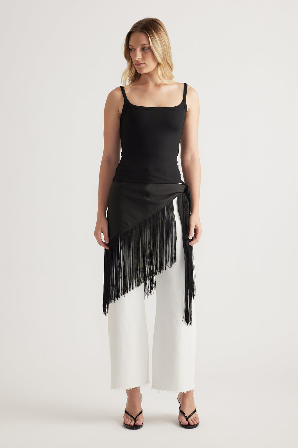 Woman wearing a black thin strap fitted tank with a fringed wrap overlay, white wide leg pants and sandals